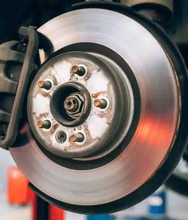 Close-up of a car brake disc and caliper, showing the shiny metal surface of the rotor and rust around the lug nut holes, with part of the wheel hub visible at the center.