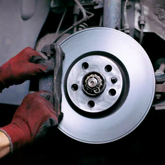 A person wearing red gloves is installing or inspecting a car's brake disc and caliper assembly, with the wheel removed to expose the brake components.